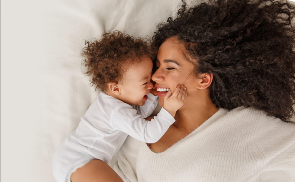Parent and baby lying on a bed, smiling and touching noses in white clothing.