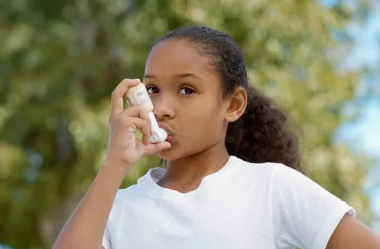 Person outdoors holding an inhaler to their mouth, wearing a white shirt.