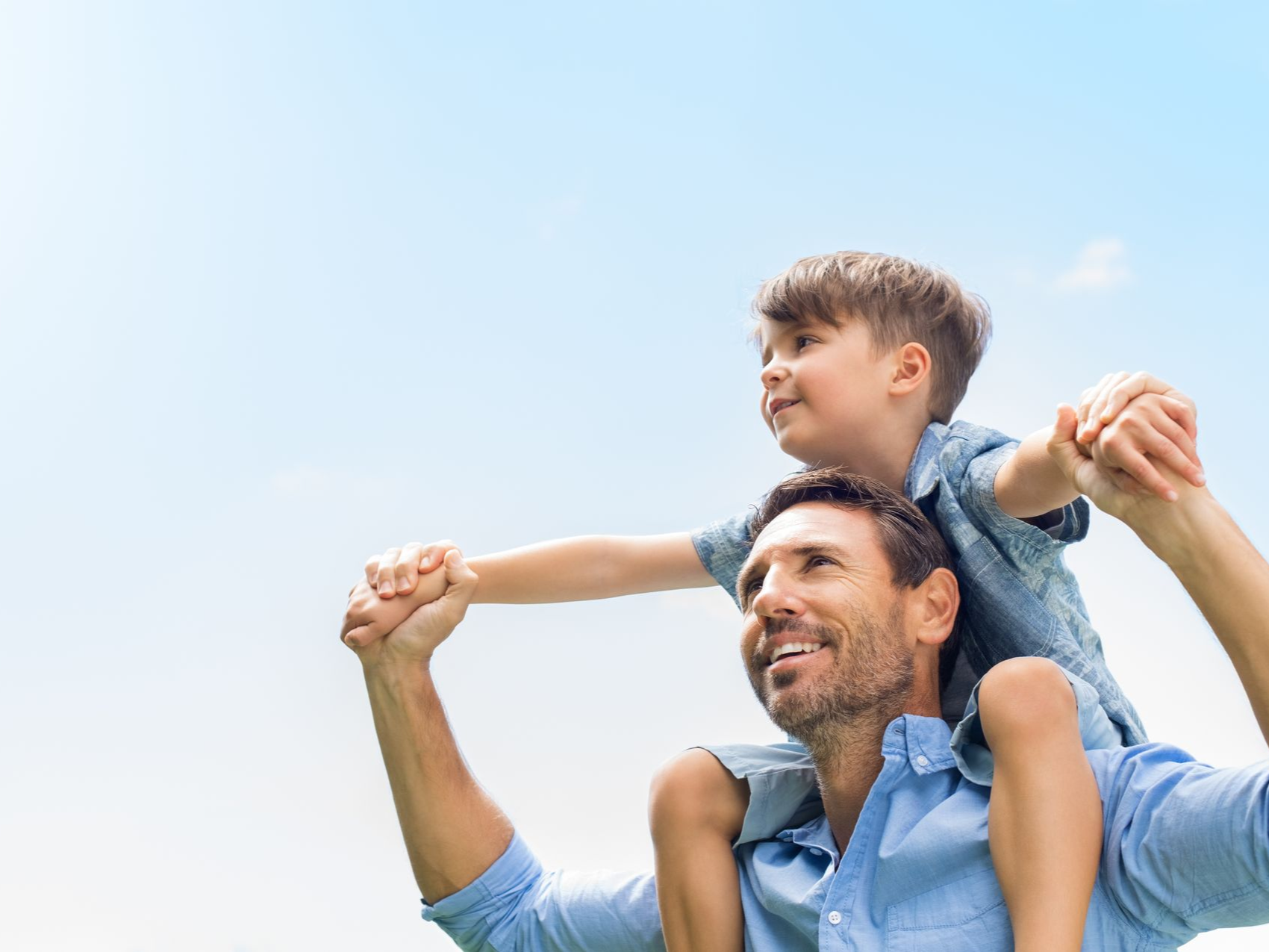 A smiling man carries a child on his shoulders against a bright blue sky.