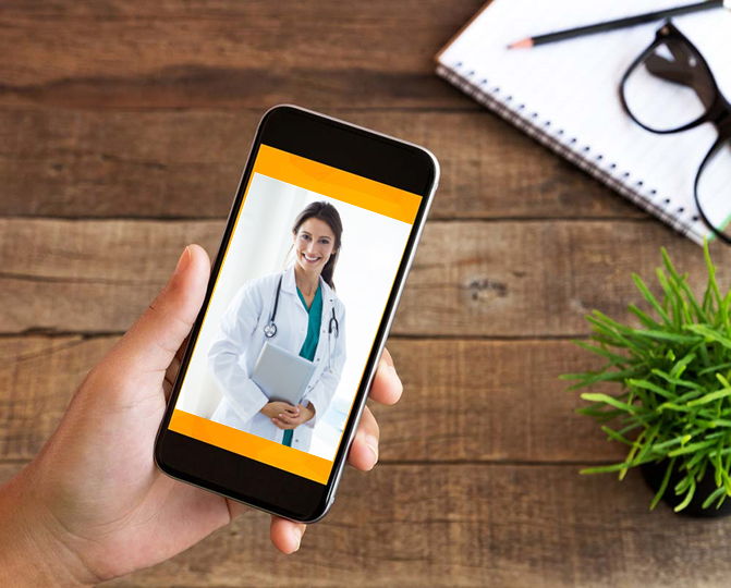 Hand holding a smartphone displaying a doctor video call on a wooden desk with notebook, glasses, and plant