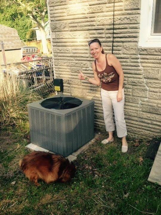 Woman smiling beside a gray outdoor AC unit, with a brown dog on the grass near a house wall.