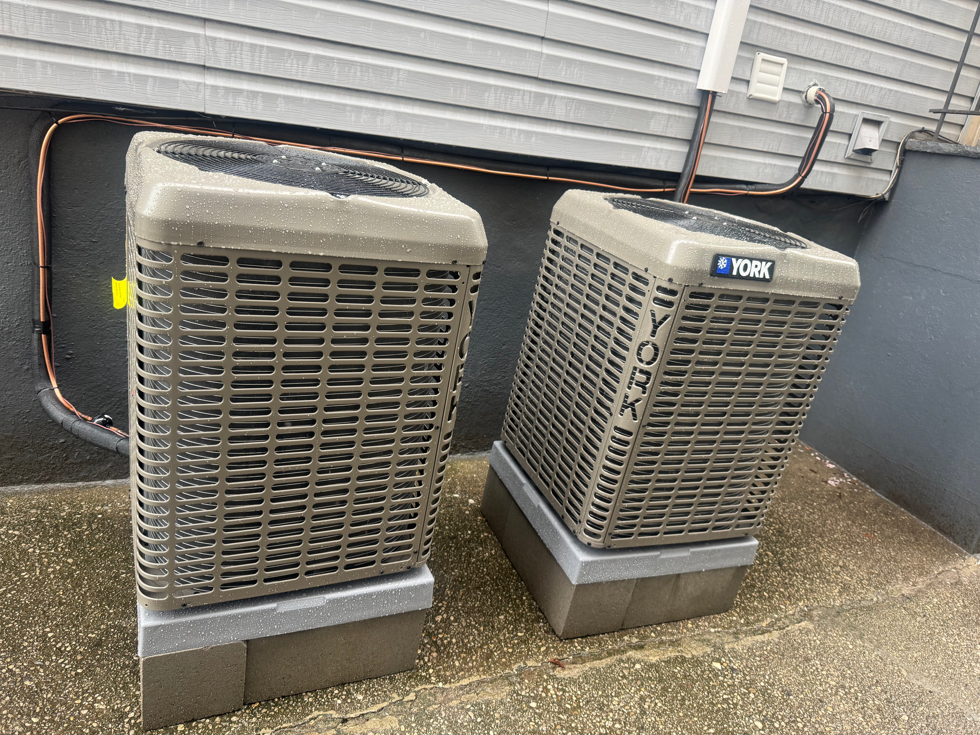 Two outdoor HVAC units beside a building wall, on concrete pads, with a hose running behind them.