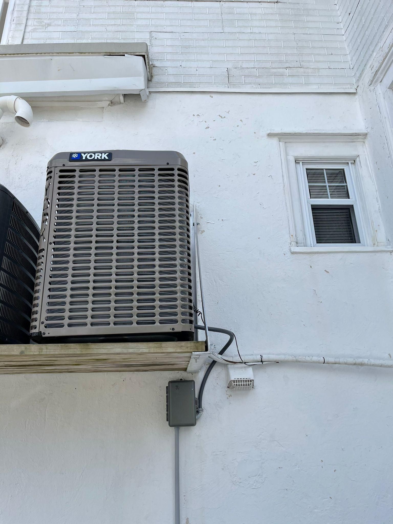 Outdoor HVAC unit mounted on a white exterior wall beside a small window and conduit box