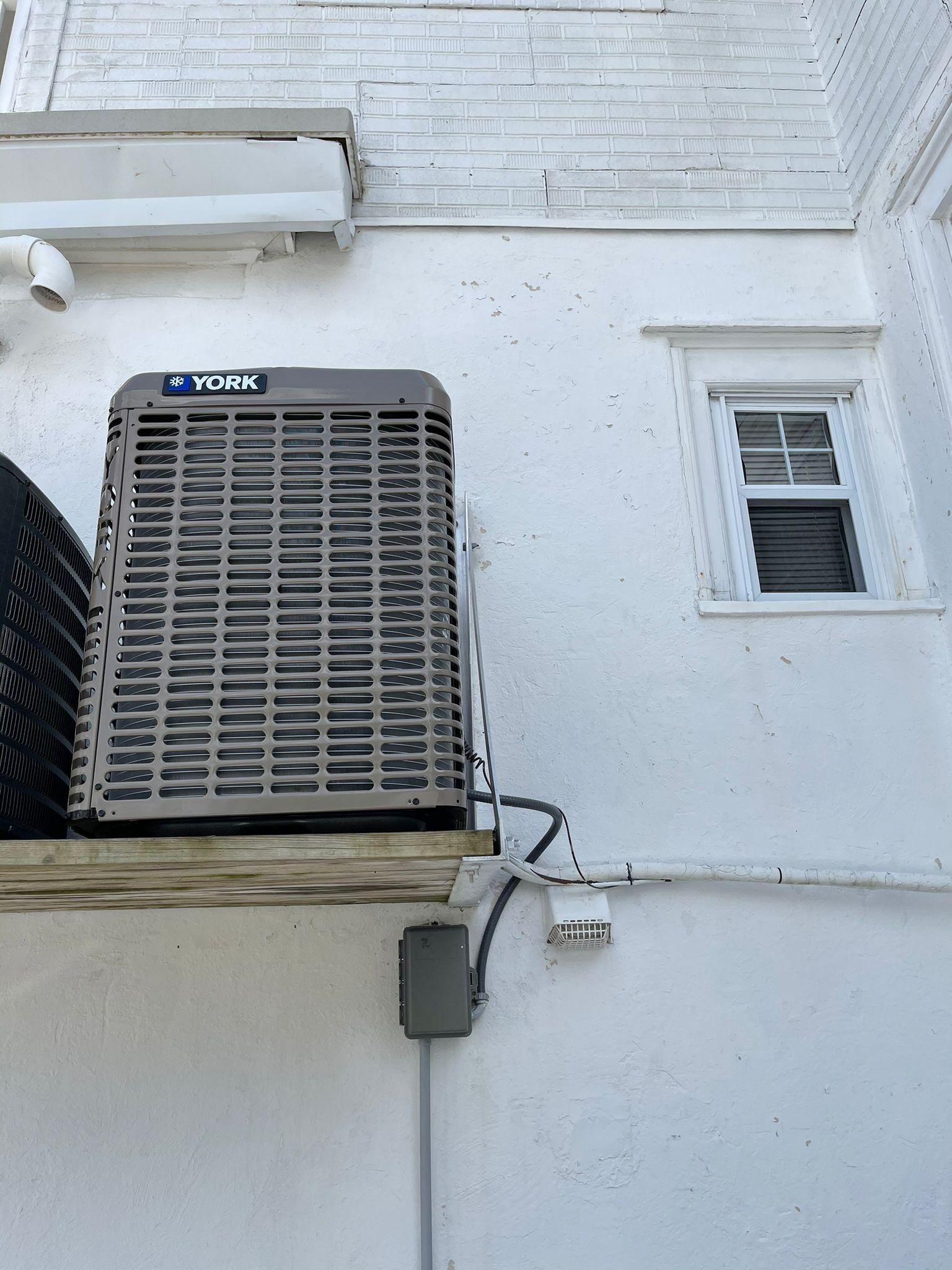 Wall-mounted York air conditioner beside a small window on a white house exterior
