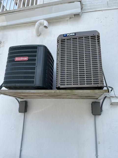 Two outdoor HVAC units mounted on a wall beneath a window, one black and one gray.