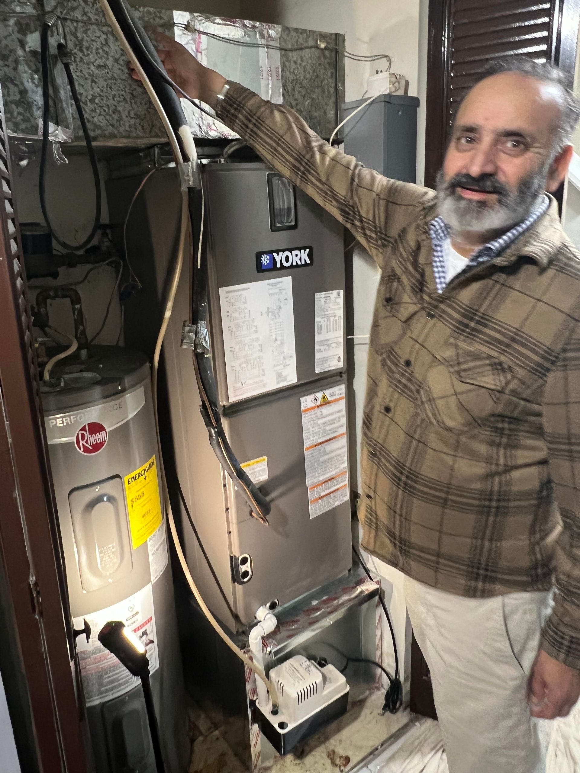 Man inspecting a furnace in a utility closet, with exposed ductwork and electrical panels nearby.