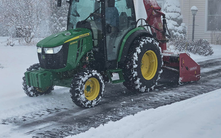 Snow blower clearing a driveway during heavy snowfall, with a red tractor and yellow wheel.
