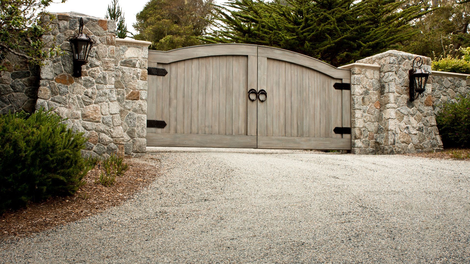 Closed wooden gate in a stone wall, with gravel driveway and trees behind it.