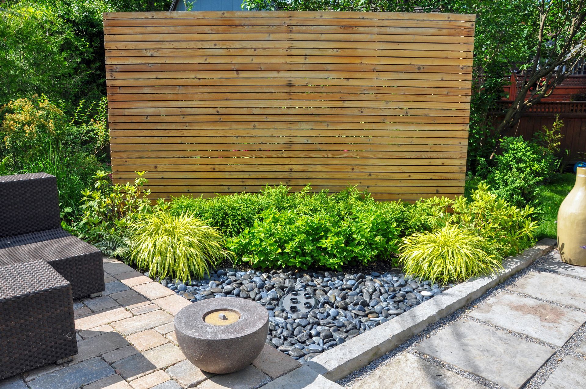 Wooden privacy screen behind lush green plants in a landscaped garden with stone pavers and pebbles
