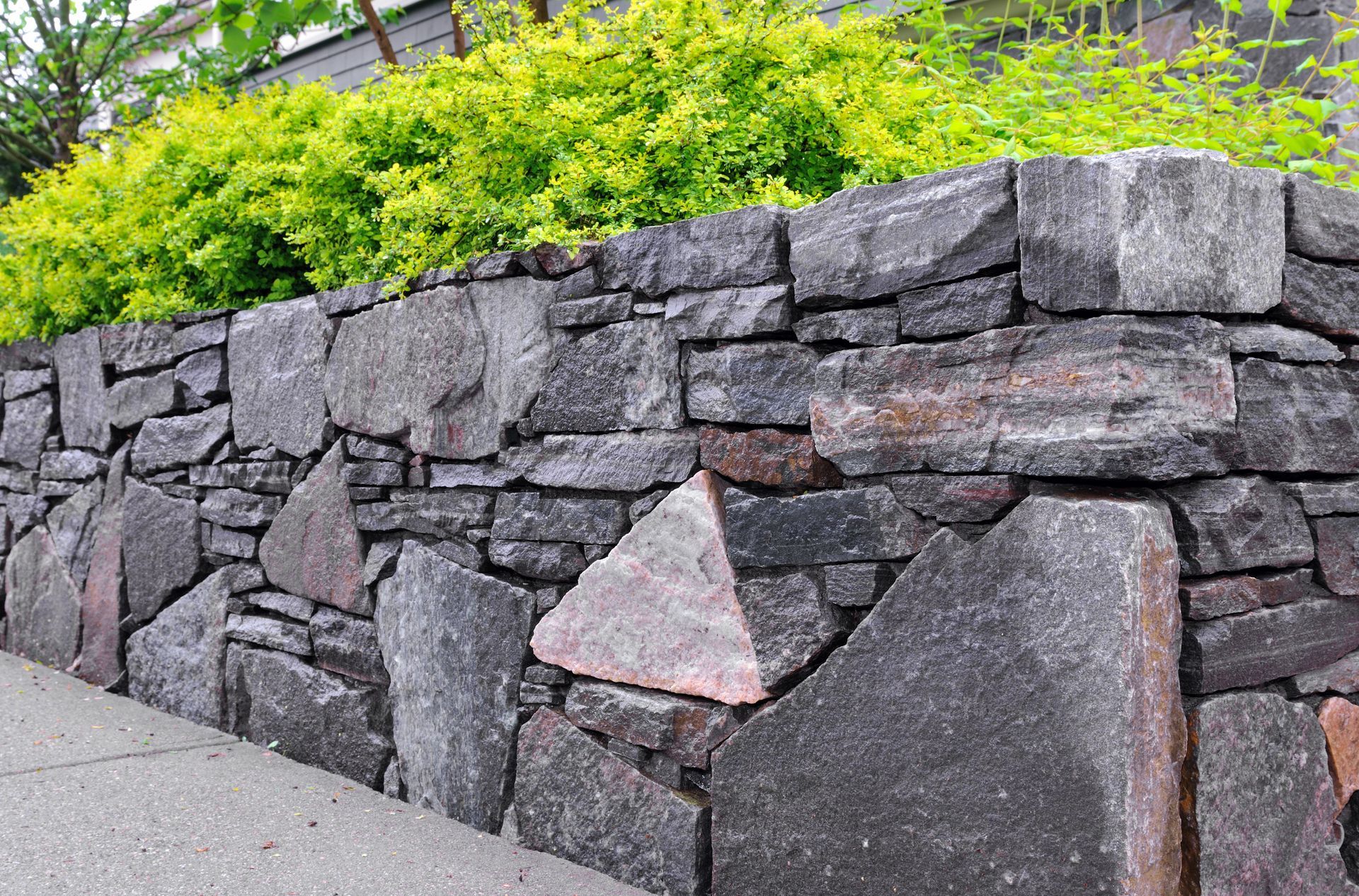 Stone retaining wall along a sidewalk, with green shrubs above and a pinkish stone accent.