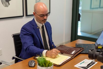 Man in a blue suit writing at a desk in an office, with paperwork, plants, and a camera nearby.