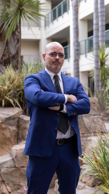 Man in a blue suit and tie standing with arms crossed in a courtyard with palm plants and buildings behind him