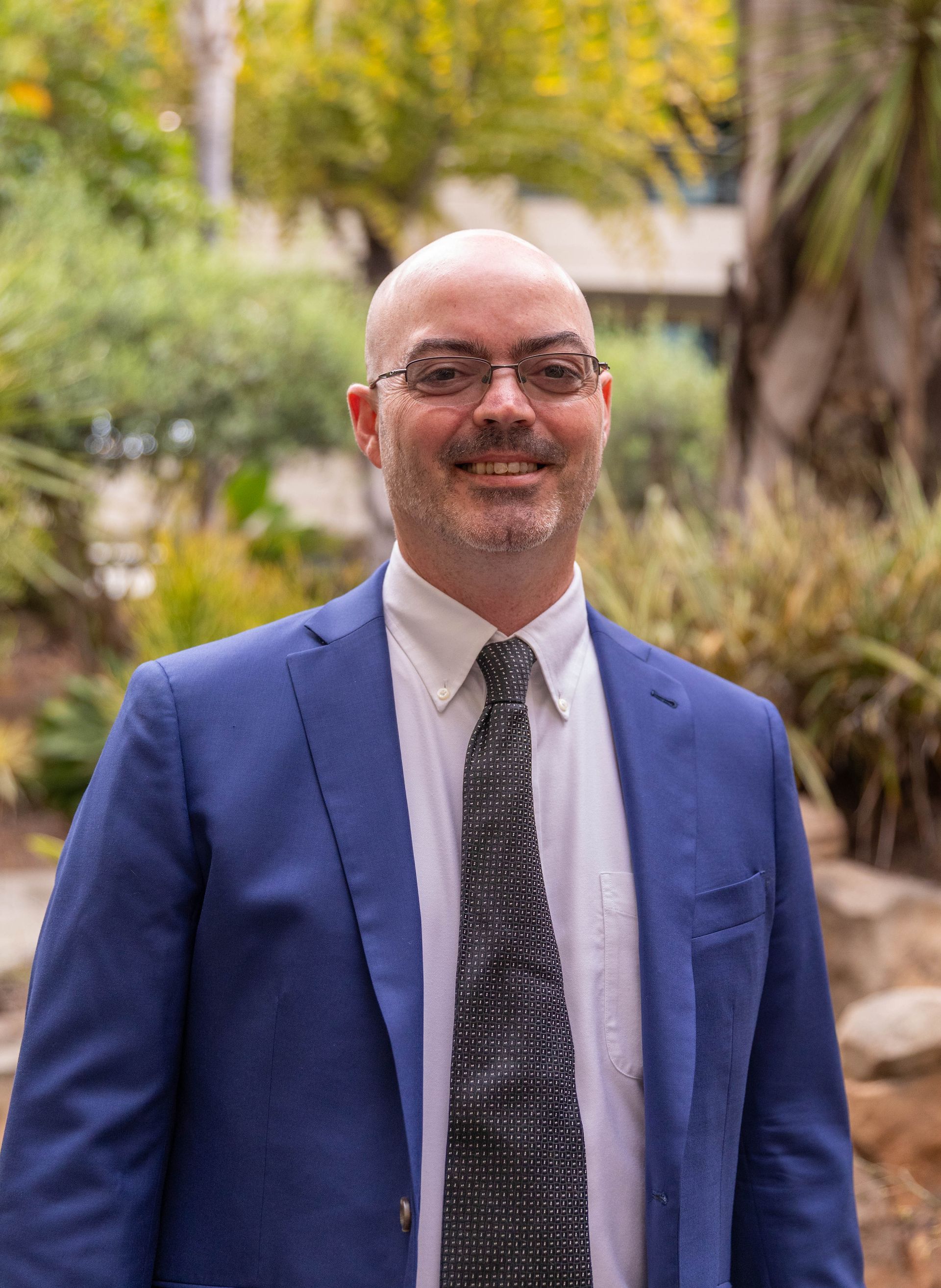 Smiling bald man in a blue blazer and tie standing outdoors in a garden.