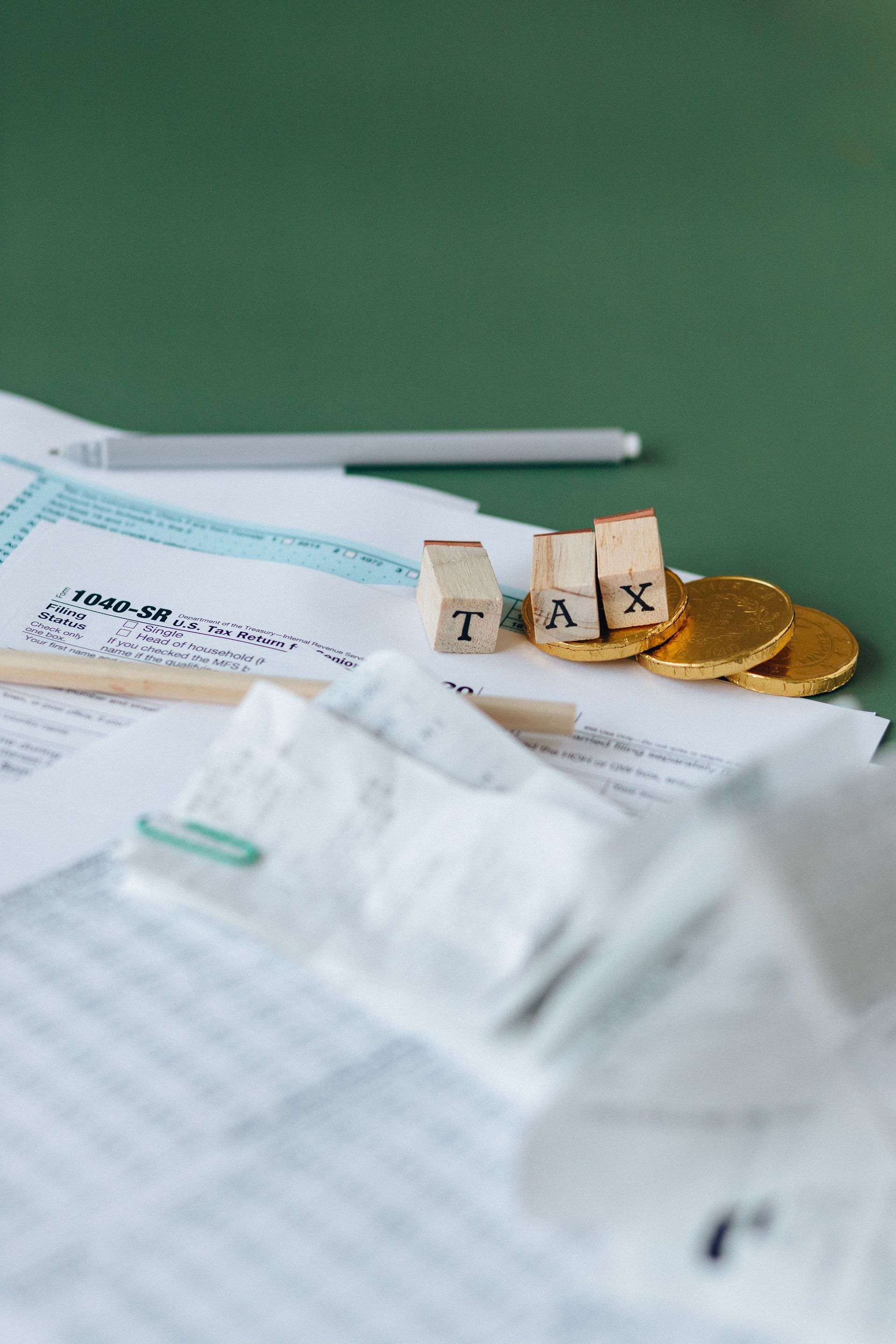 Tax forms with a pen and wooden “TAX” blocks on a desk with coins.