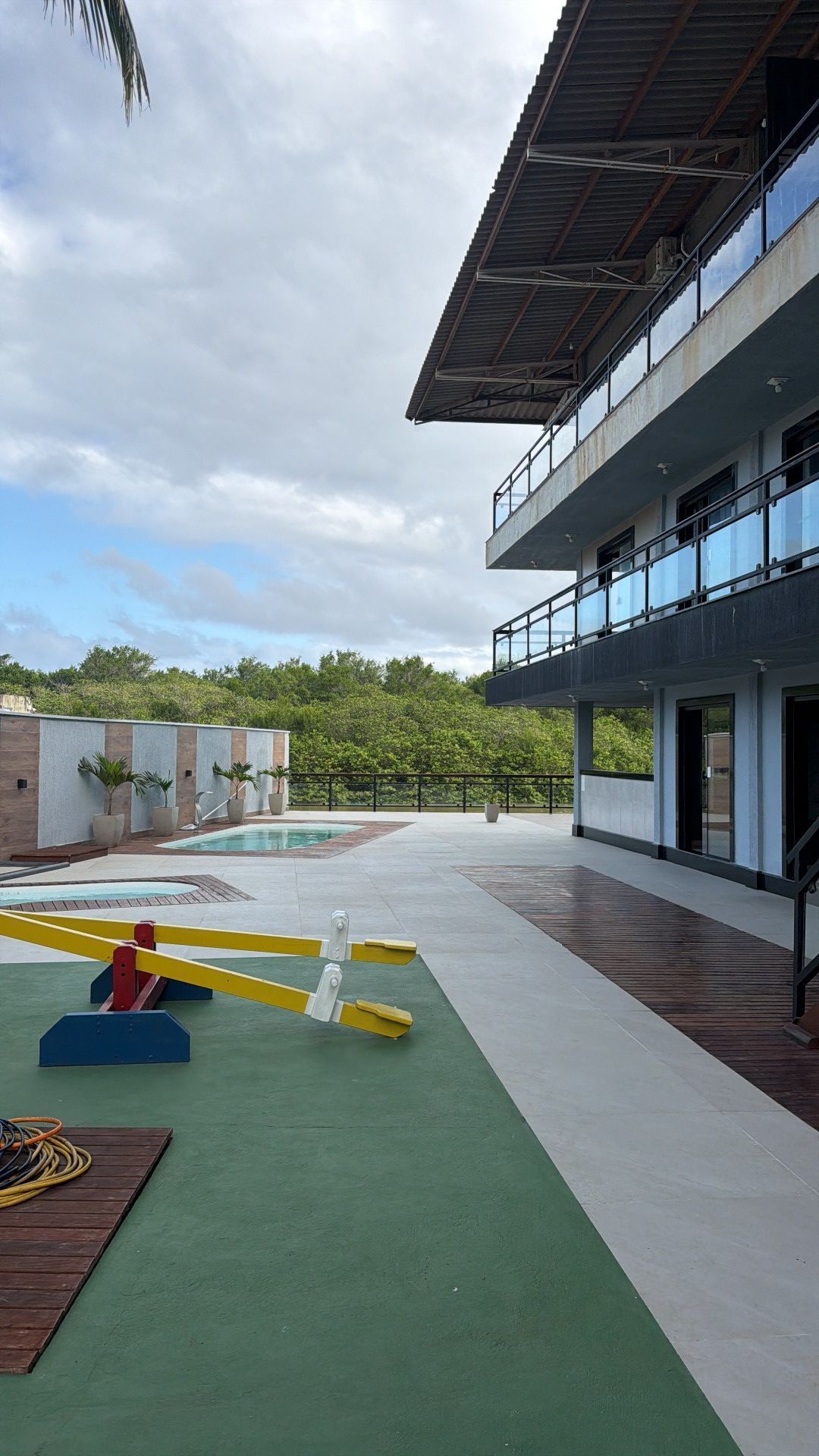 Pátio externo vazio ao lado de um edifício moderno, com um brinquedo de parque infantil sobre um piso verde e céu nublado.