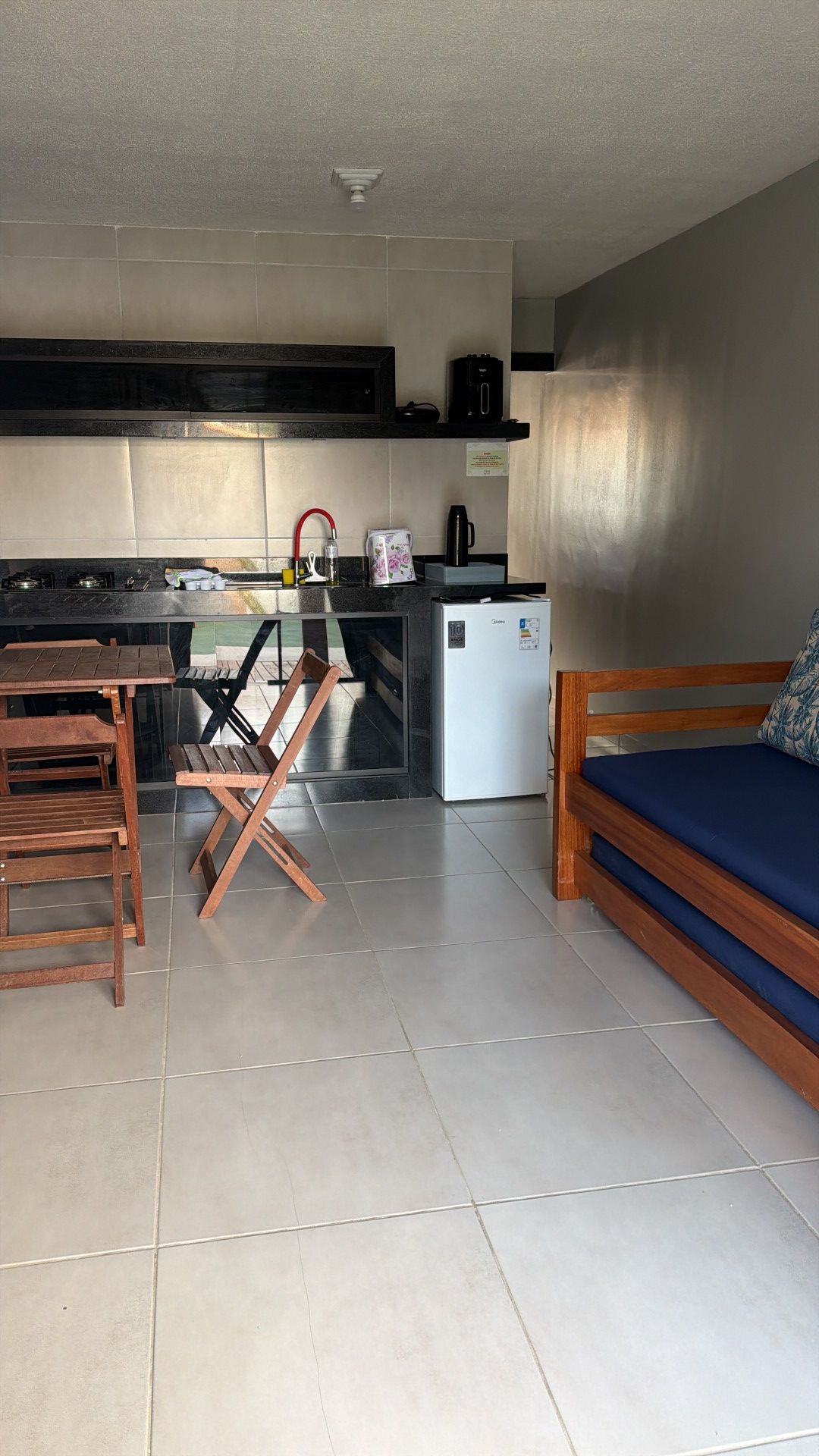 Estúdio pequeno com cozinha, mesa dobrável e cadeiras, sofá azul e piso de azulejos.
