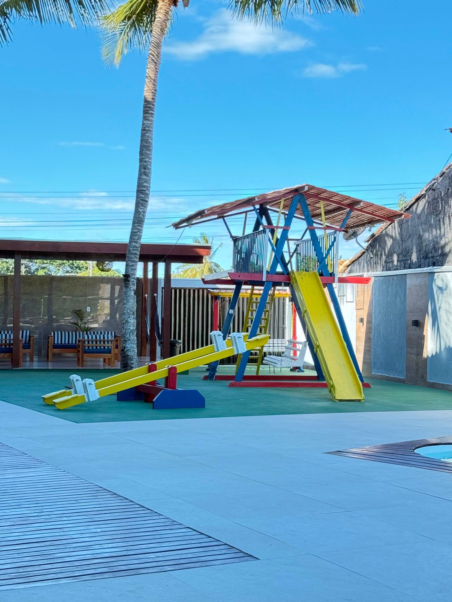 Parque infantil com escorregas coloridos e estrutura de escalada junto a uma zona de descanso à sombra de palmeiras