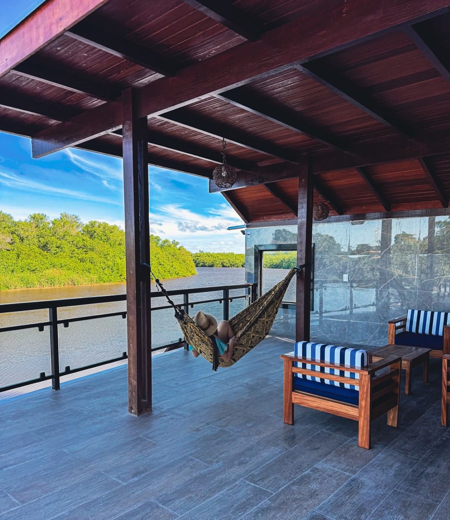 Deck coberto com uma cadeira de rede, mesa e cadeiras de madeira, com vista para um rio e uma margem verdejante.