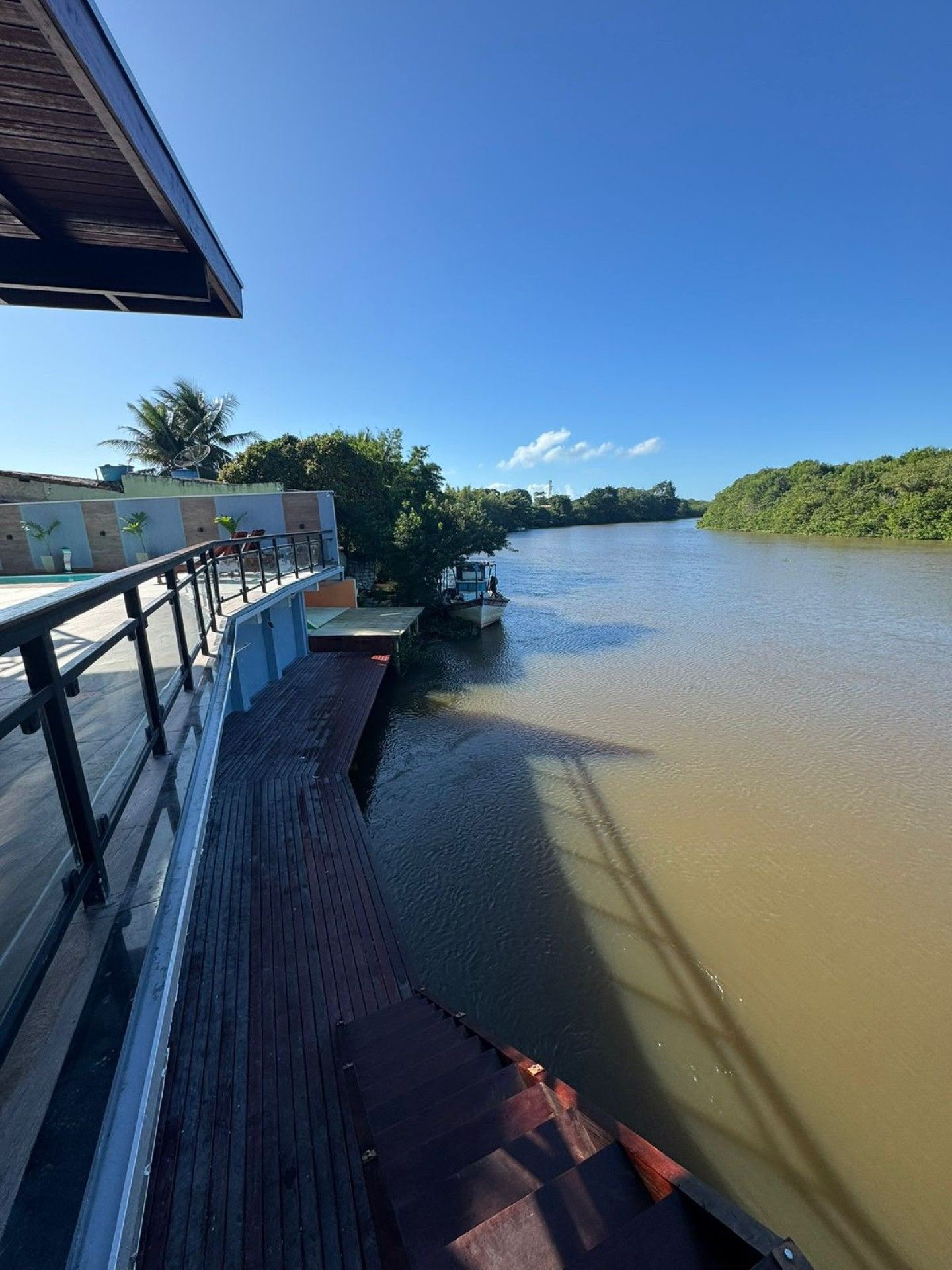 Calçadão à beira-rio com grade de proteção, águas tranquilas e marrons, margem verde e céu azul-claro brilhante
