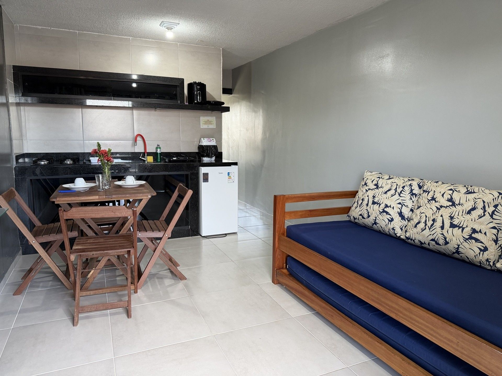 Sala de estar e jantar pequena, com uma mesa de madeira, uma área de cozinha e um sofá com almofadas azuis junto à parede
