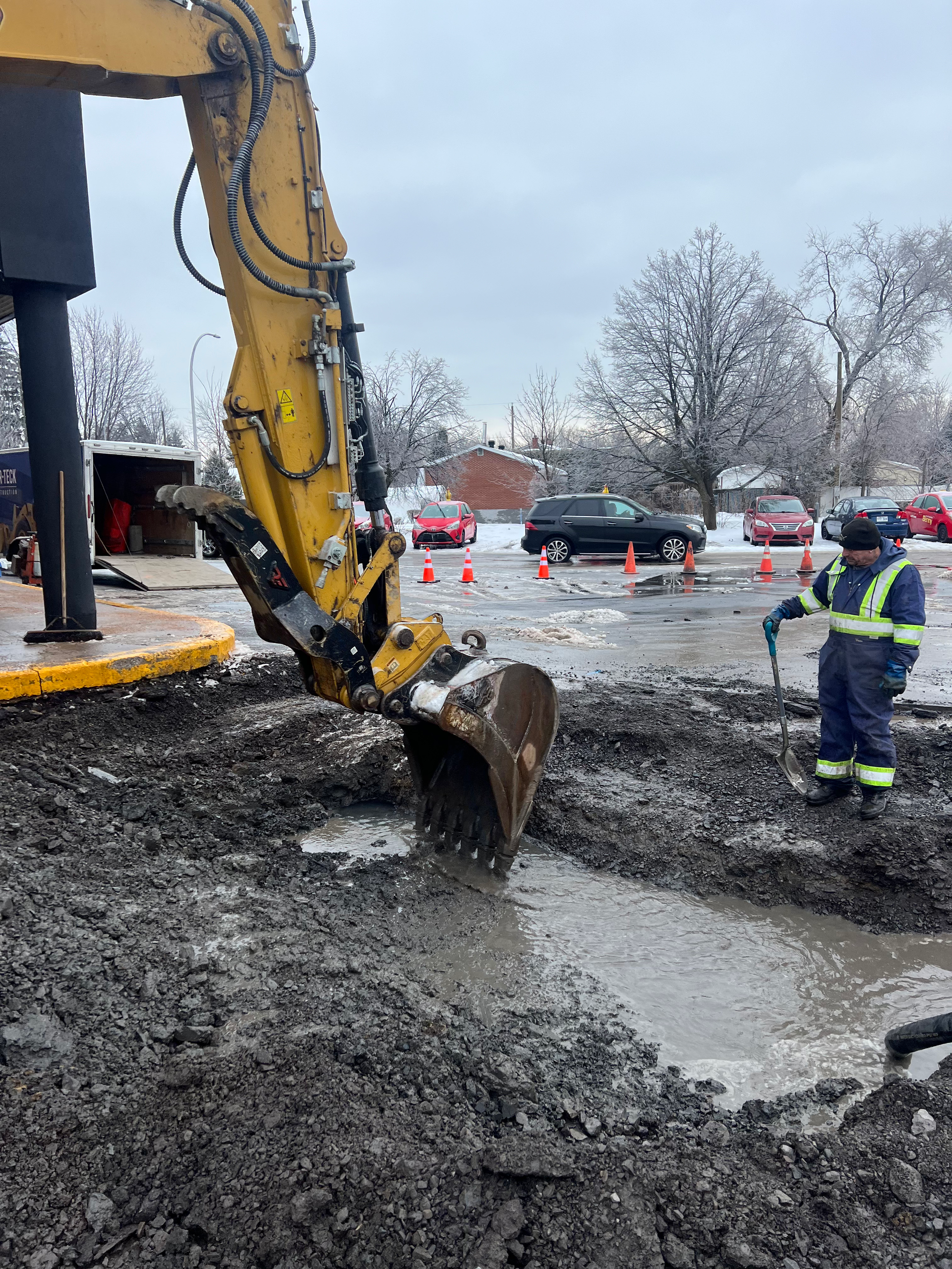 Excavator digging in muddy ground next to a worker in high-visibility clothing. Cloudy, outdoor setting.