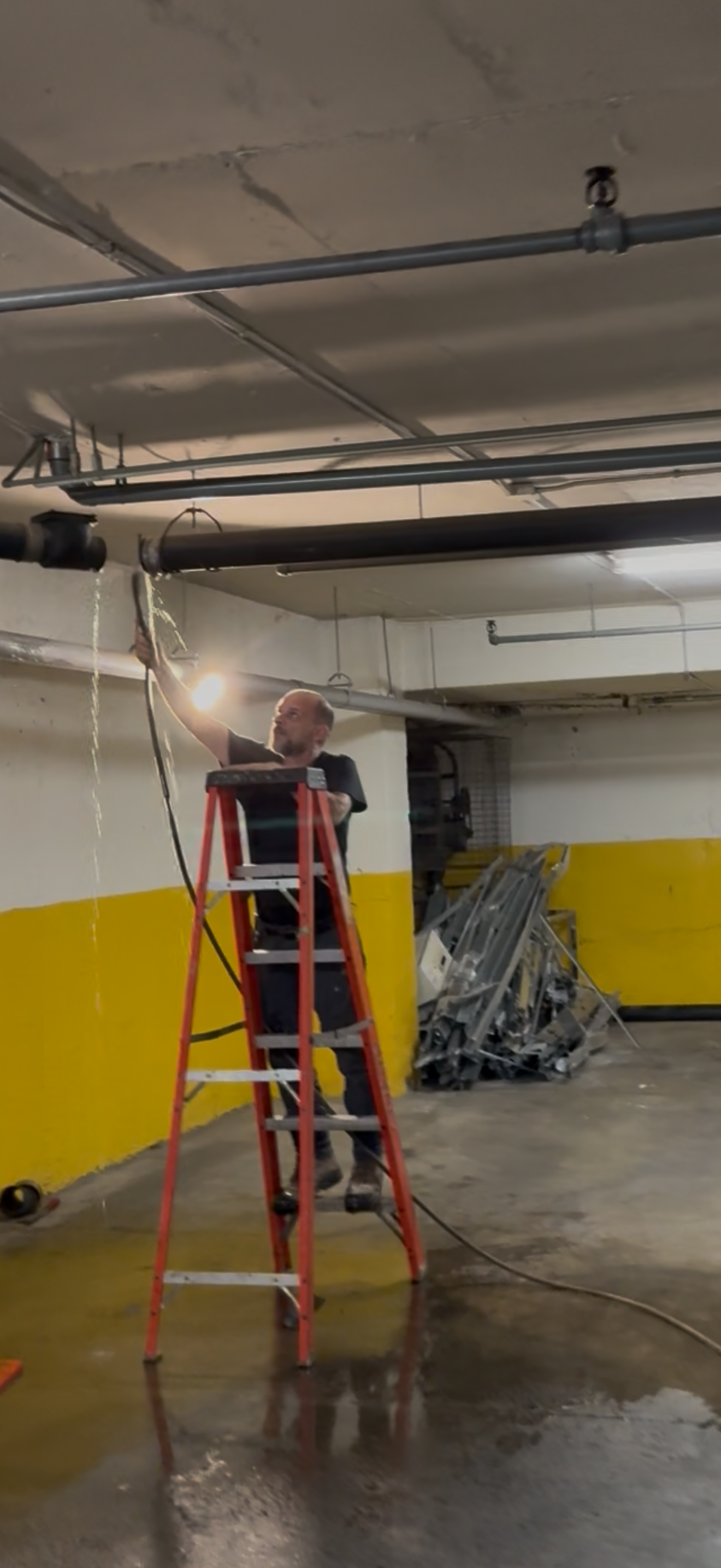 Person on a ladder working on pipes in a garage. Sparks fly. Yellow and concrete walls.