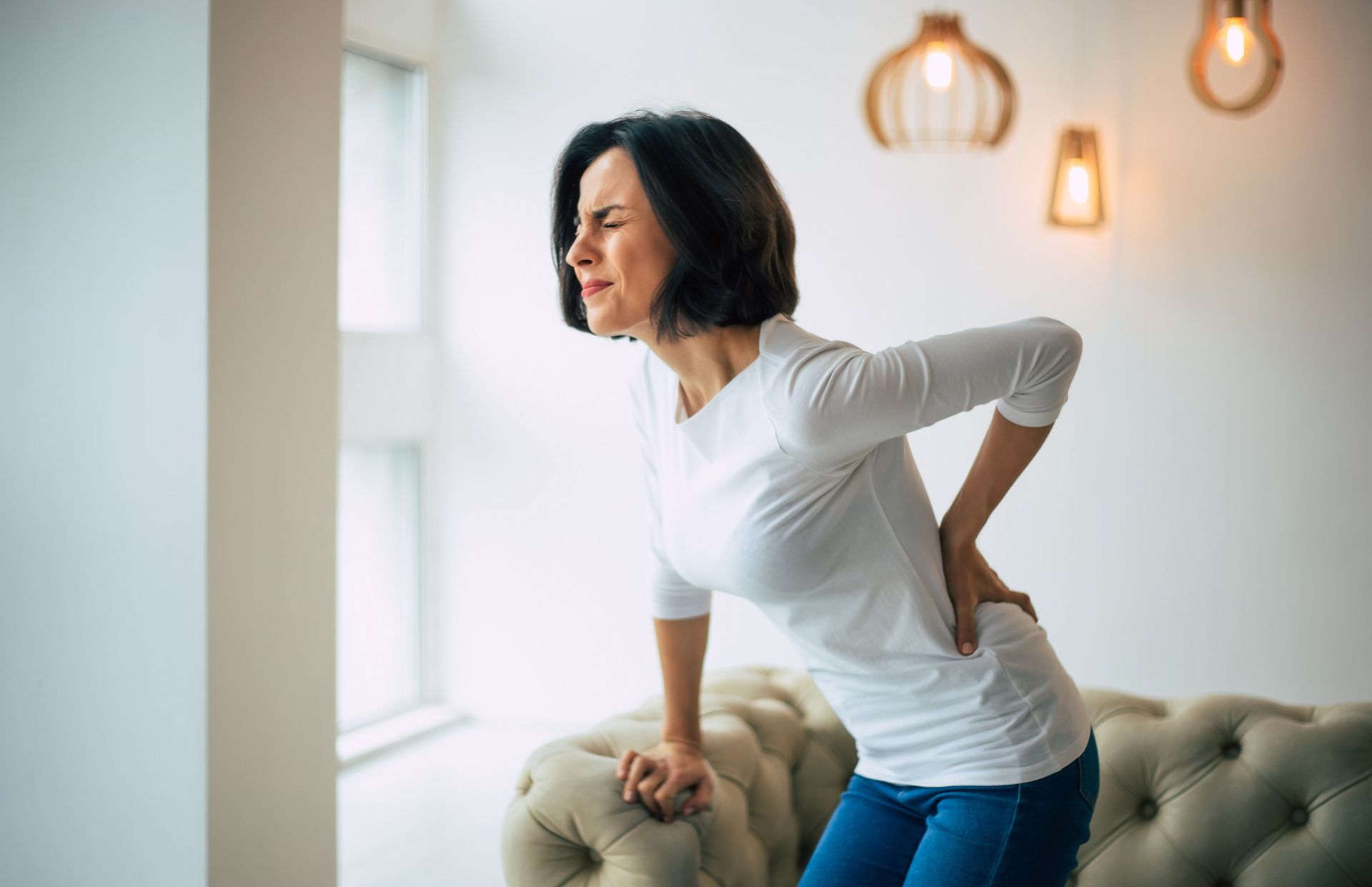 A woman is sitting on a couch holding her back in pain.