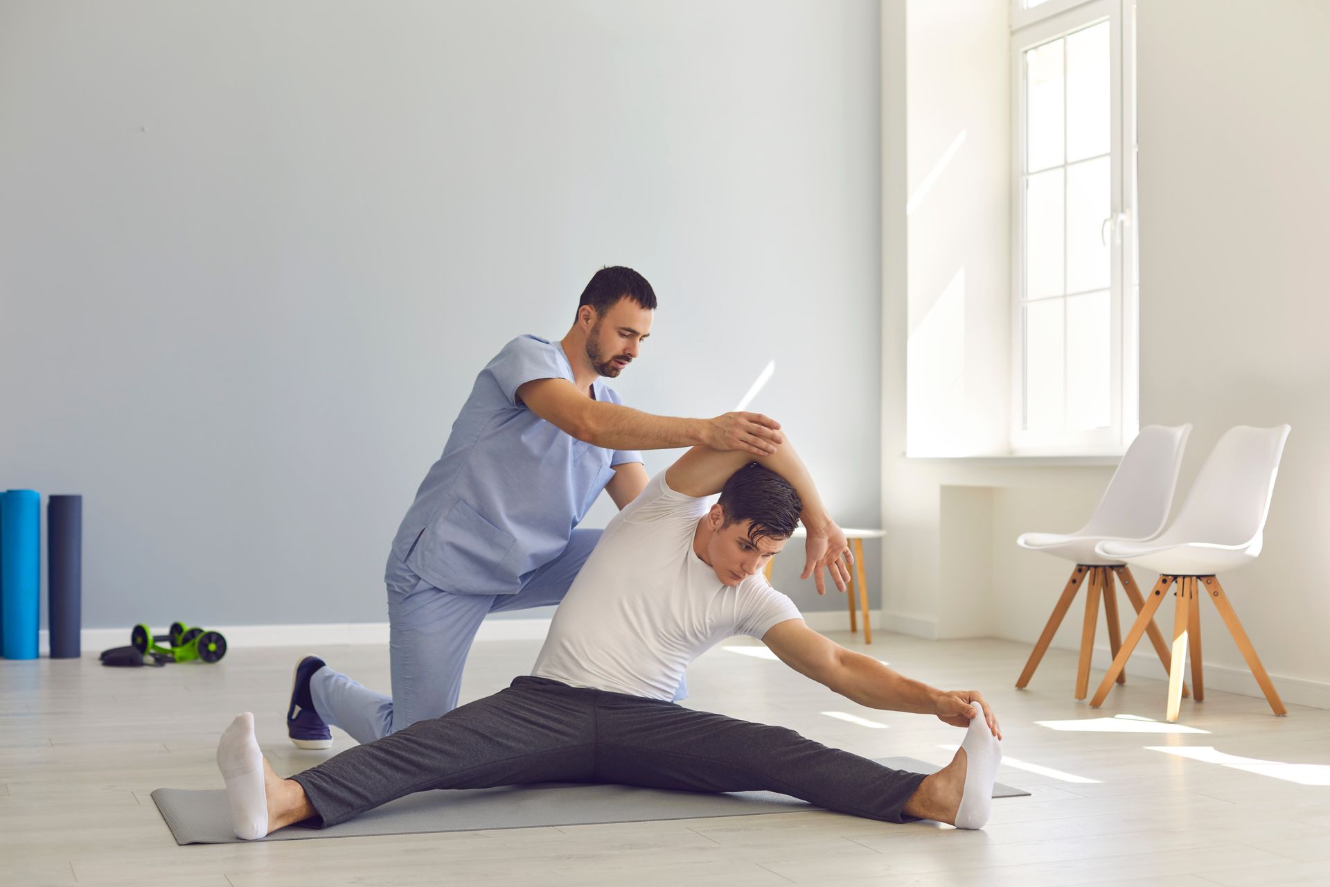 A man is sitting on the floor stretching his legs while a nurse helps him.