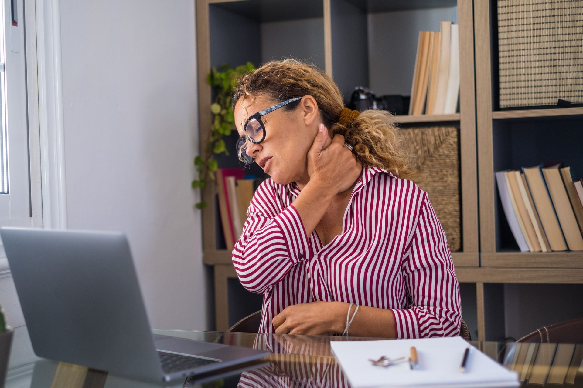 A woman is sitting at a desk with a laptop and holding her neck.