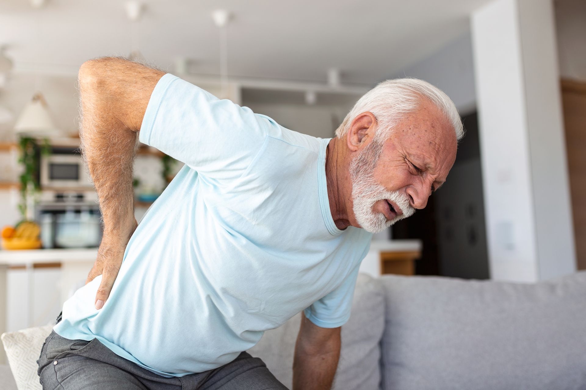 An elderly man is sitting on a couch holding his back in pain.