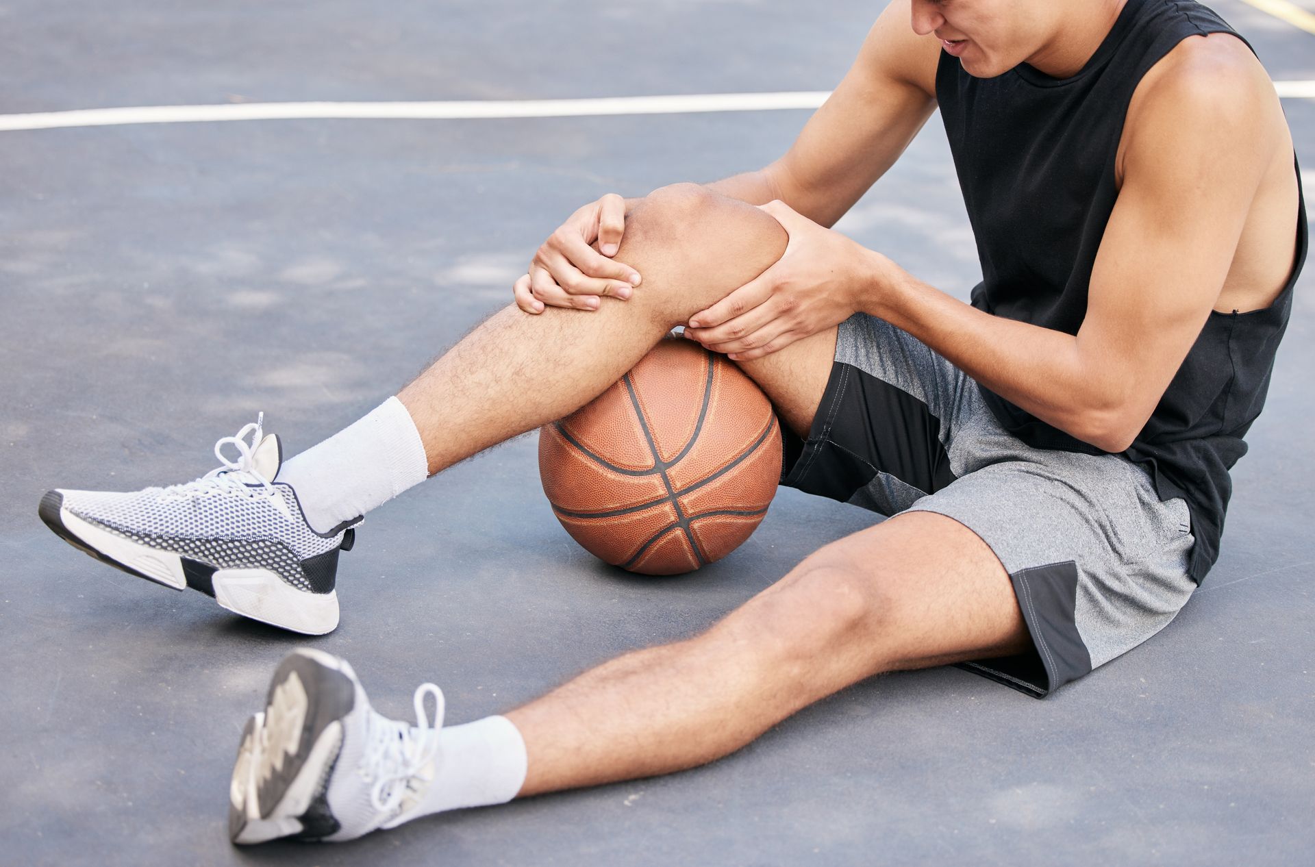 A man is sitting on the ground holding his knee while holding a basketball.
