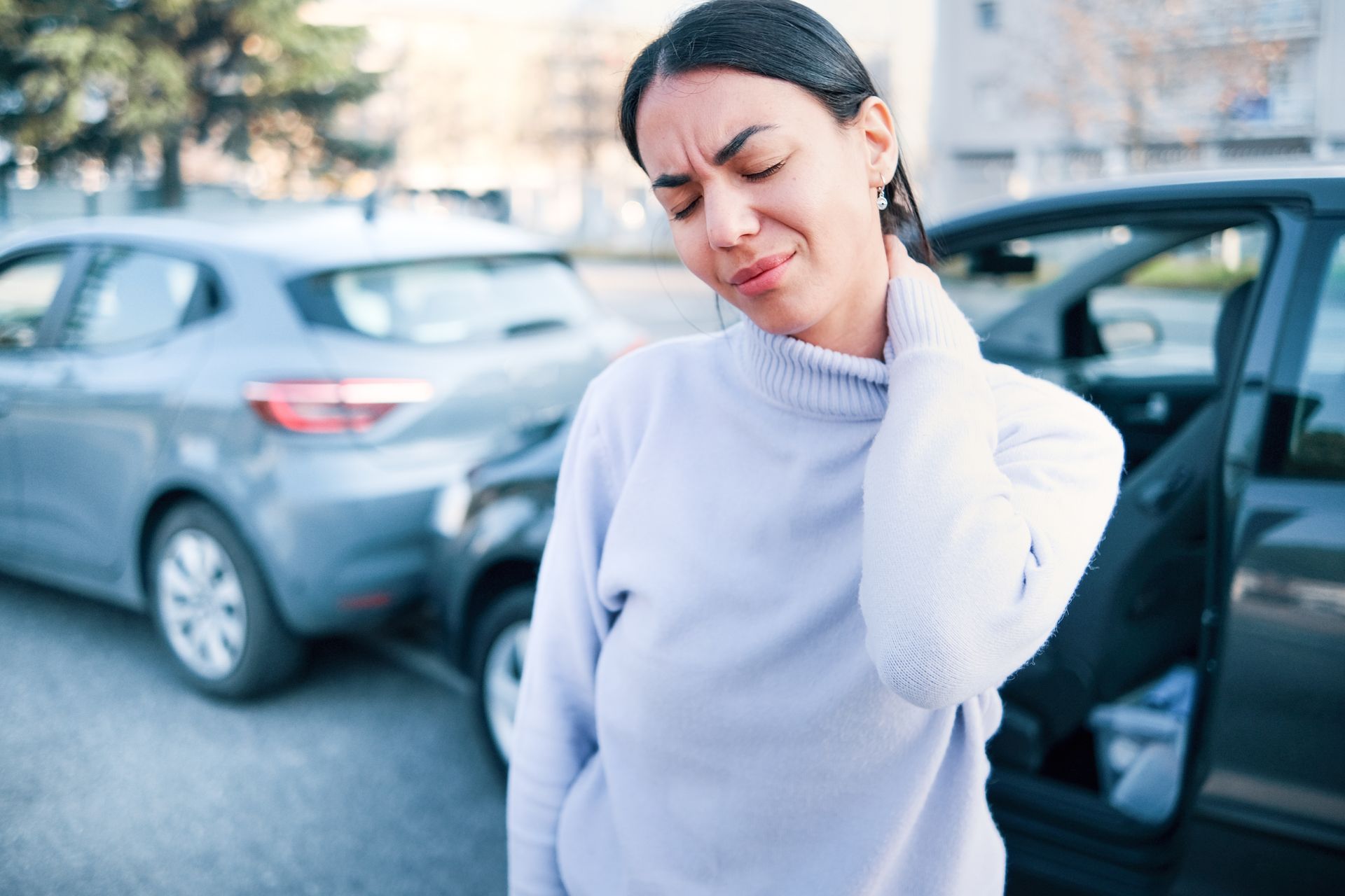 A woman is holding her neck in pain in front of a car.