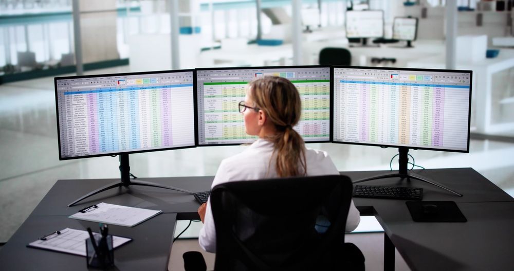 Woman seated at desk, working on three computer monitors displaying spreadsheets in an office setting.