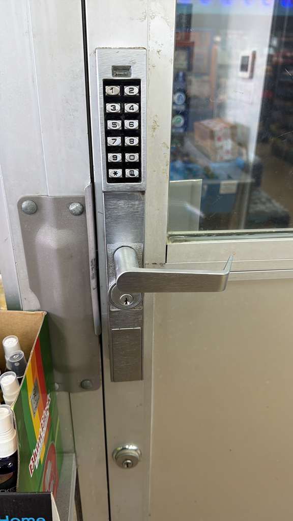 Metal door with keypad, handle, and a keyhole. The door is closed, with a box of products visible on the left.