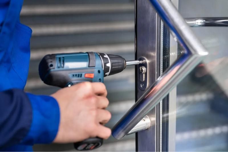 Person in blue overalls using a power drill to install a door handle on a metal door.