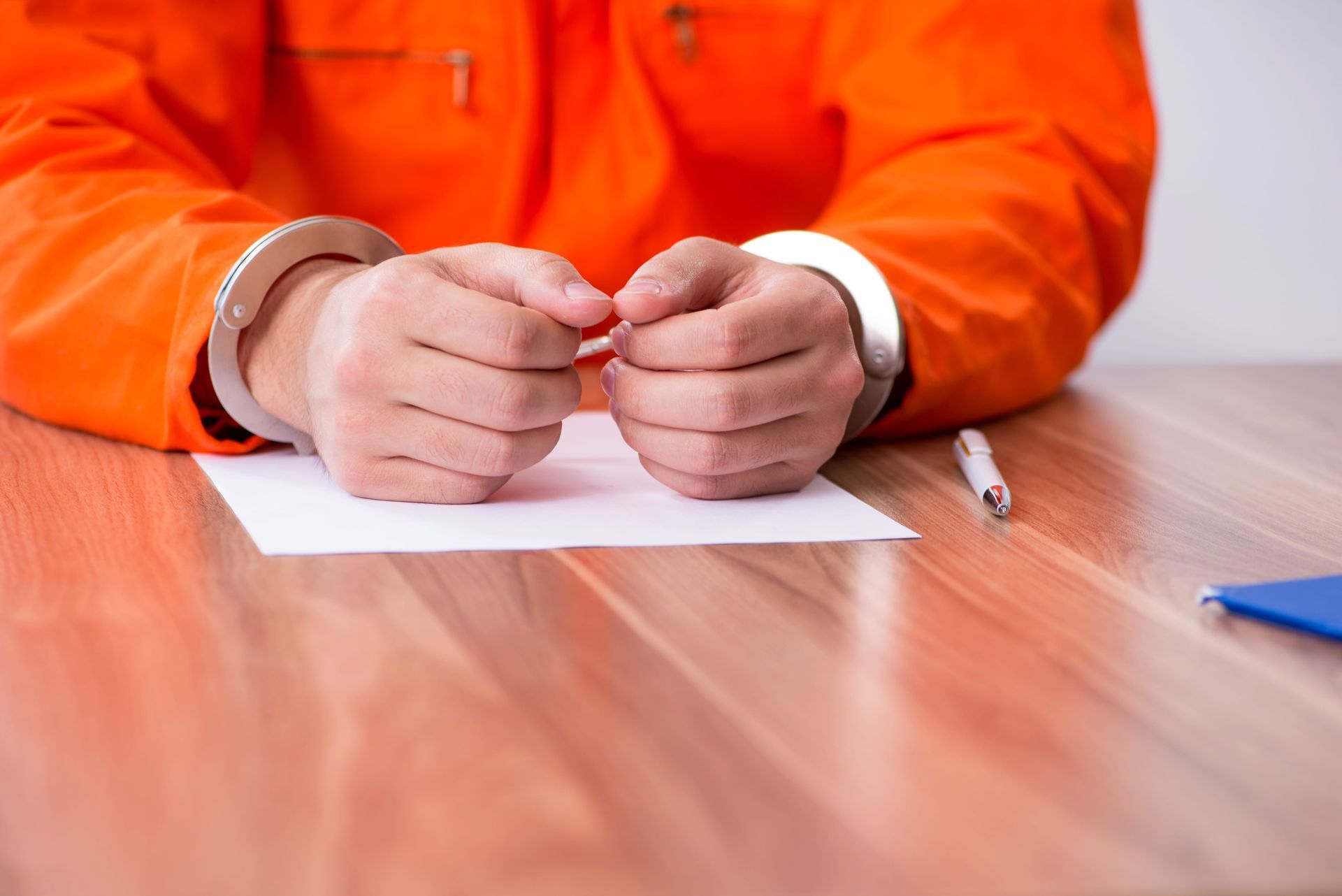 Person in an orange jacket with handcuffs sits at a table, hands clasped over a sheet of paper.