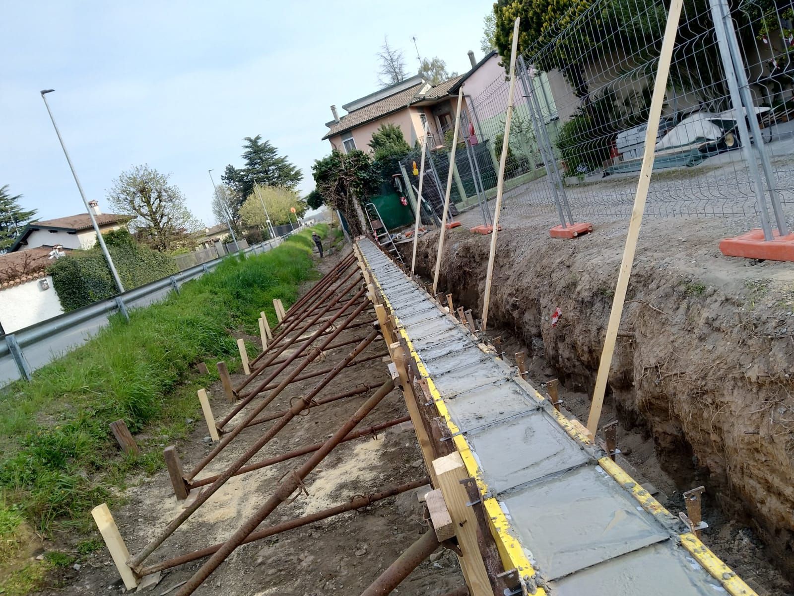 Scavo di fondazione in cemento in corso di realizzazione, rivestito con casseforme gialle e sostenuto da pali metallici angolati in un'area erbosa.