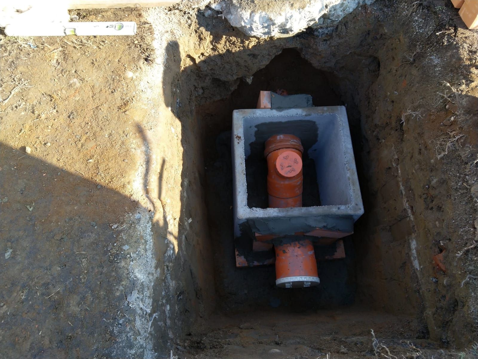 Una camera di ispezione rettangolare in cemento per il drenaggio è ricavata in una buca scavata nel terreno e collegata a tubi in PVC arancioni.