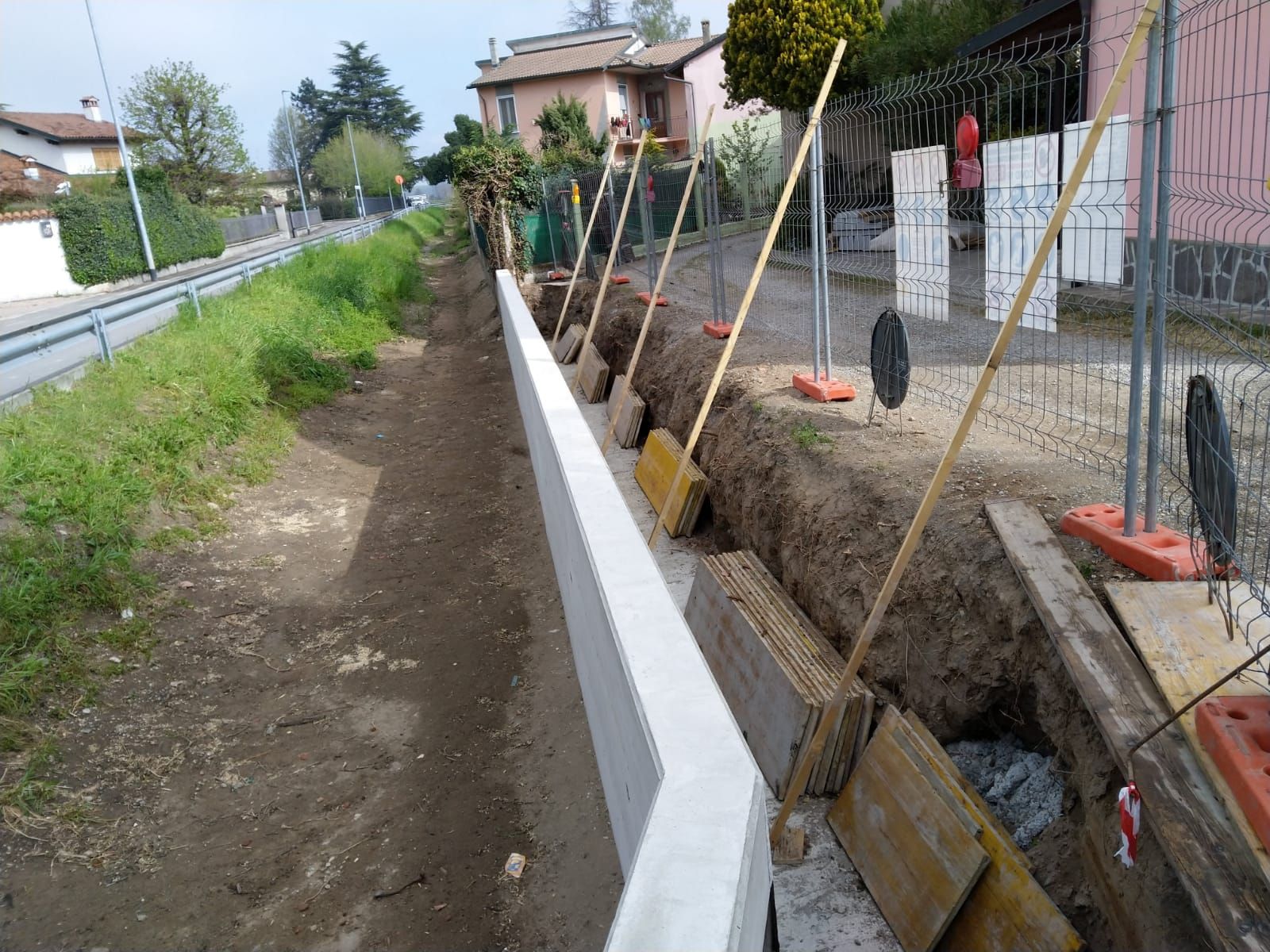 Un muro di cemento in costruzione accanto a uno scavo di terra, delimitato da una recinzione metallica provvisoria da cantiere.