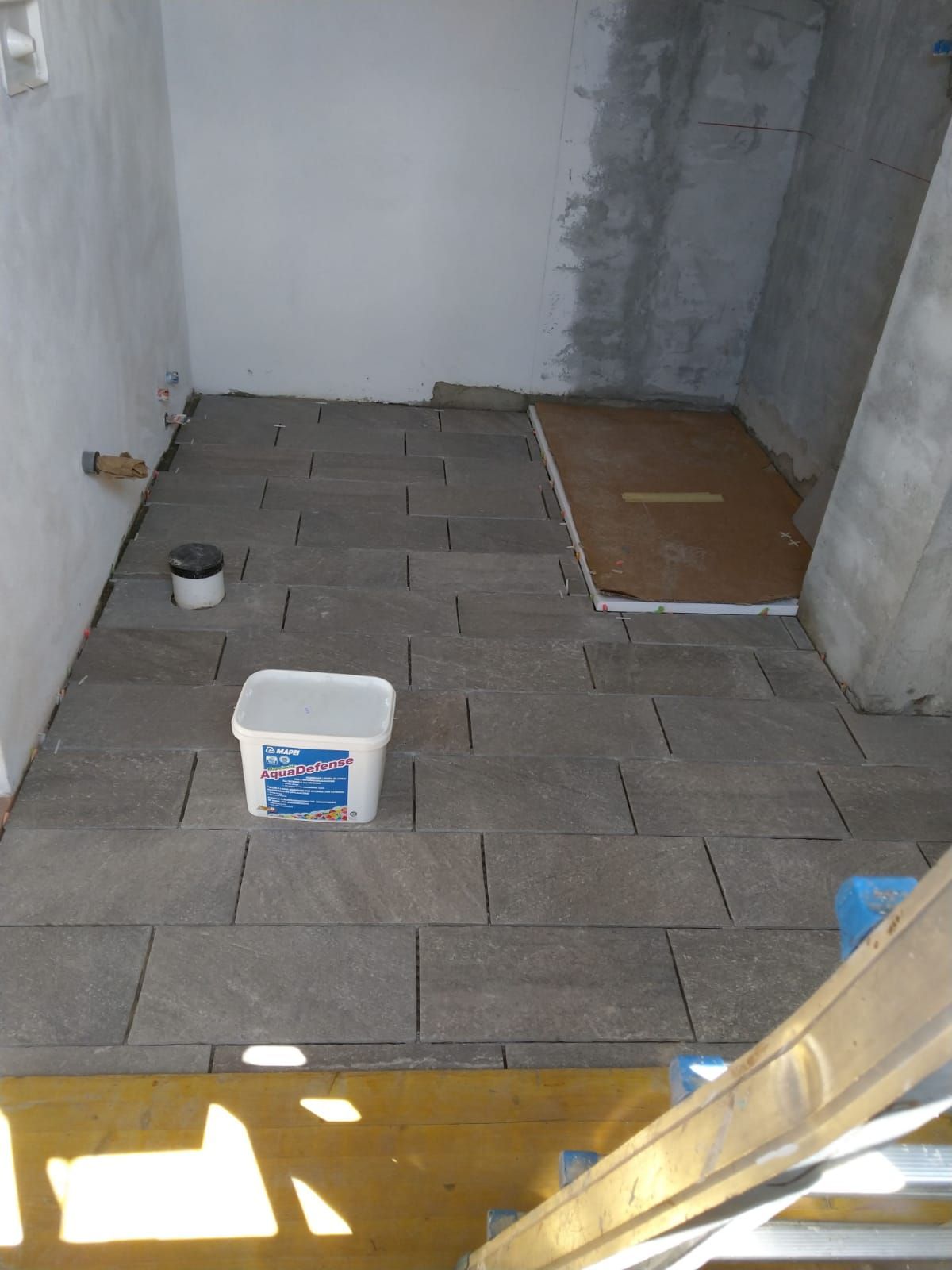 Una stanza in costruzione con pavimento piastrellato in piastrelle rettangolari grigie, un secchio e una zona doccia protetta.