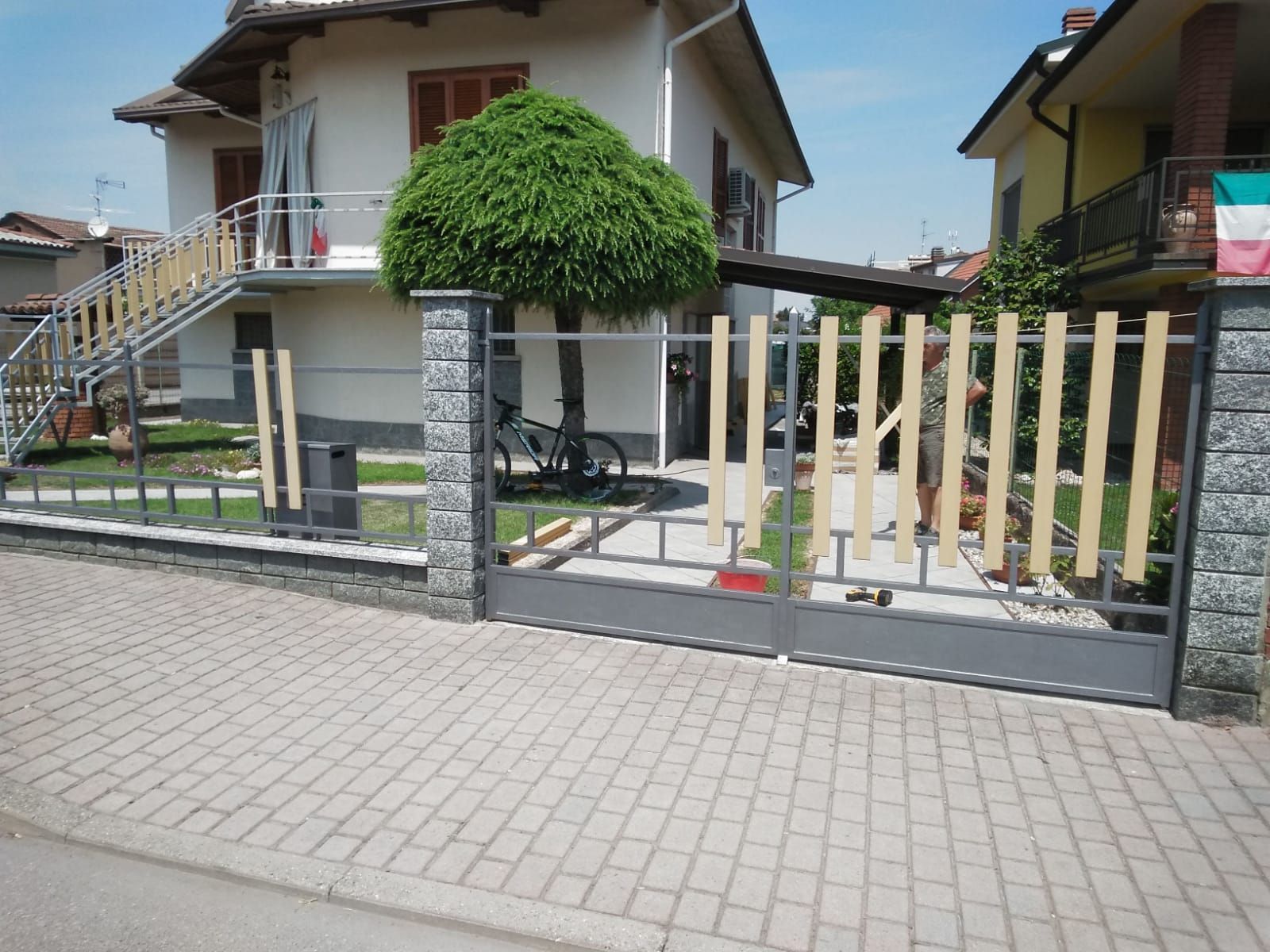 Una casa residenziale con un moderno cancello in metallo grigio caratterizzato da lamelle verticali di colore chiaro, situato dietro un marciapiede pavimentato.