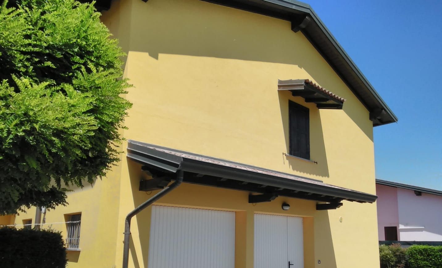 Una casa gialla a due piani con tetto a due falde scuro, porte del garage bianche, una piccola tenda da sole alla finestra e un grande albero sulla sinistra.