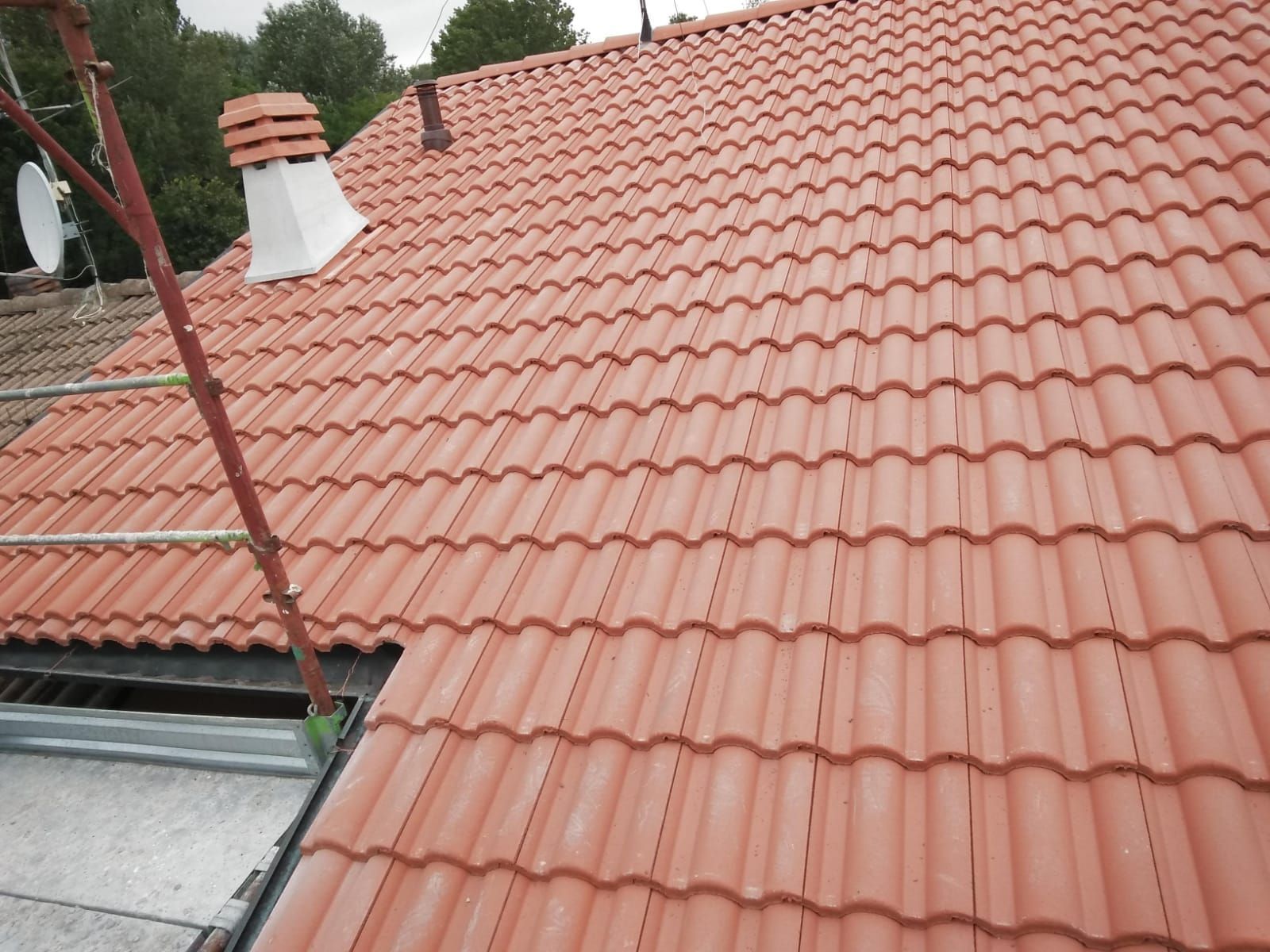 Un tetto spiovente ricoperto di tegole di terracotta rossa con un camino, un tubo di ventilazione e un'impalcatura nelle vicinanze.