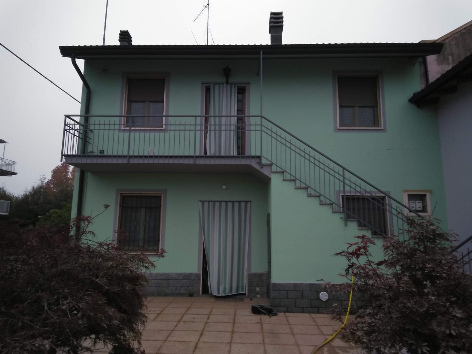 Una casa a due piani, di colore verde chiaro, con un patio piastrellato, una scala esterna in metallo e un balcone, in una giornata nuvolosa.