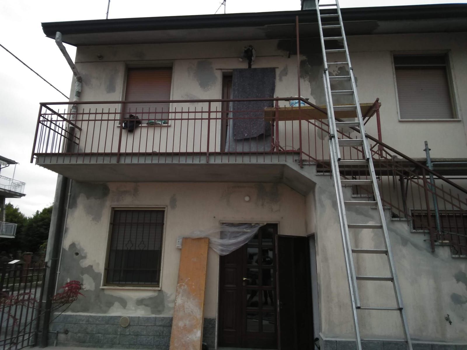 Esterno di una casa a due piani in fase di ristrutturazione, con balcone, porta aperta, tavola di legno e scala appoggiata su di essa.
