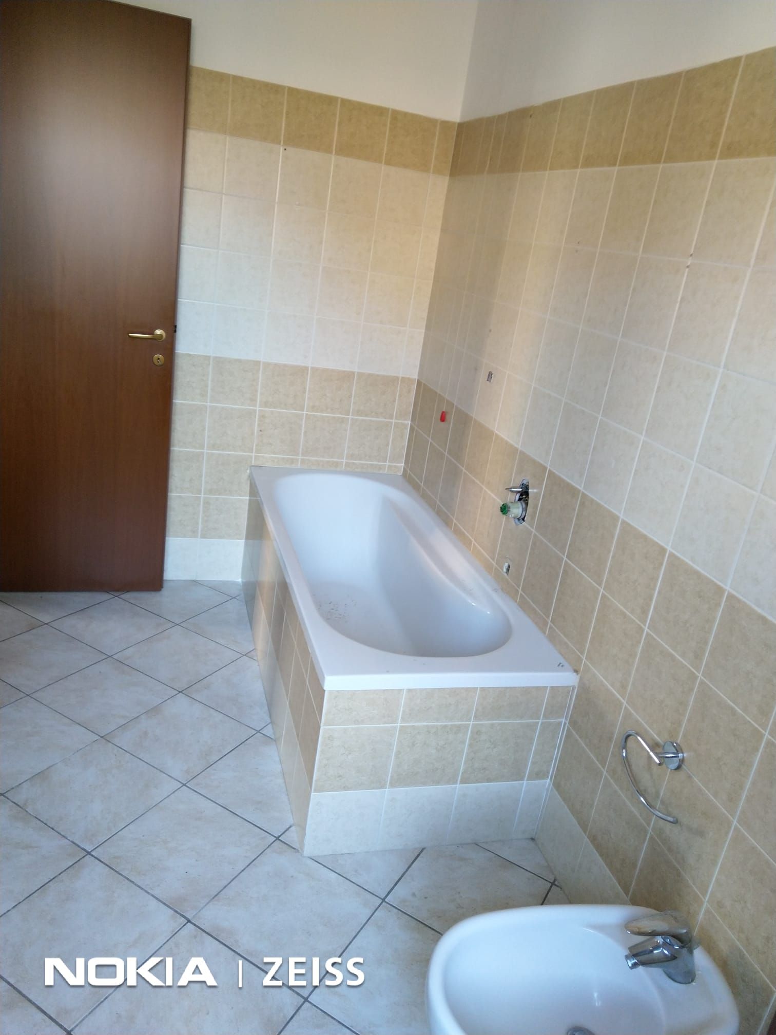 Un bagno con vasca bianca su pareti piastrellate beige, bidet in primo piano e porta in legno sulla sinistra.