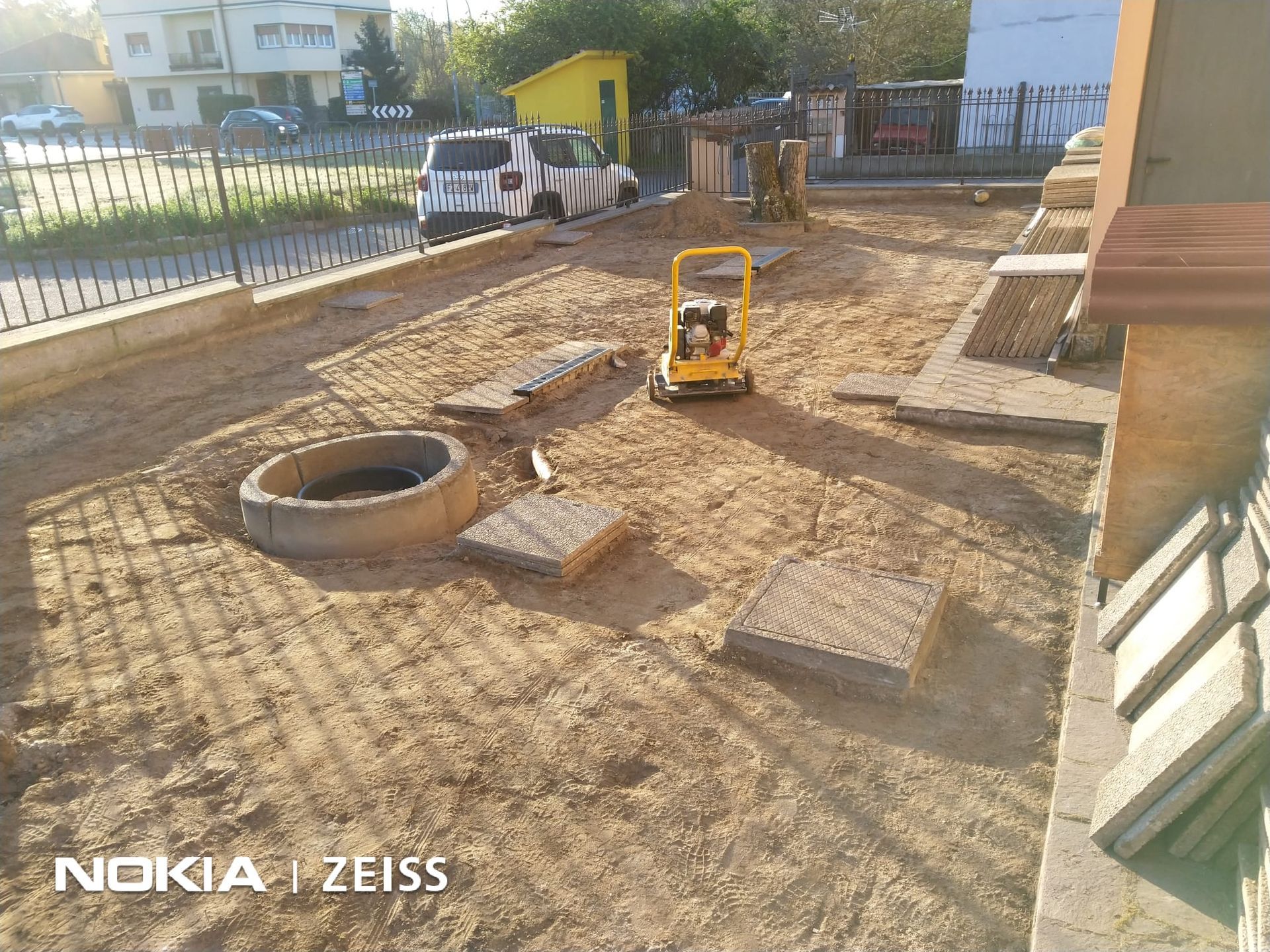 Un cantiere in un cortile con una piastra vibrante gialla, sabbia sciolta e lastre di pavimentazione vicino a un anello di cemento.
