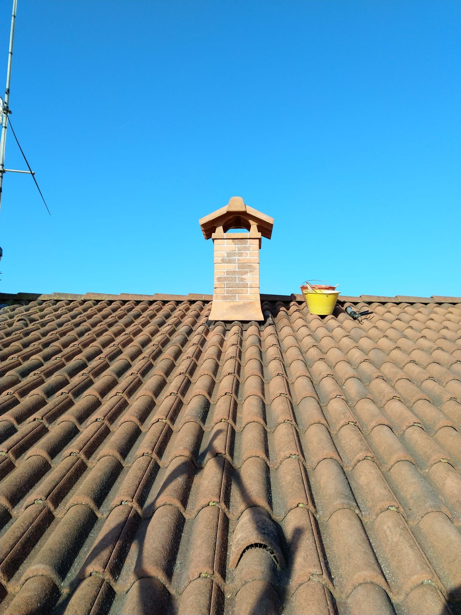 Un camino in mattoni con un piccolo tetto a punta si erge su un tetto di tegole marroni, stagliandosi contro un cielo azzurro e limpido.