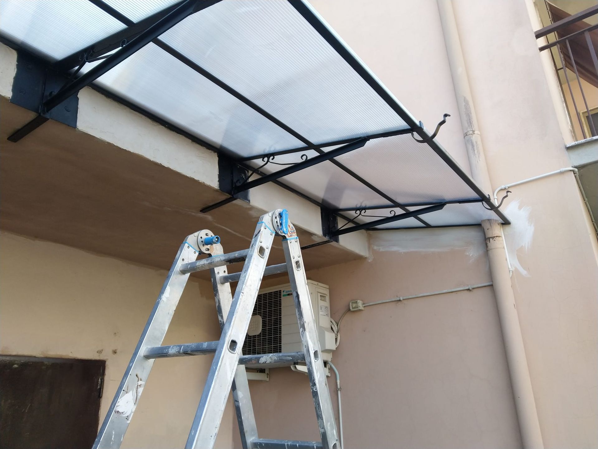 Una tenda da sole in policarbonato con struttura metallica installata su una parete esterna beige, con una scala a pioli in alluminio in primo piano.