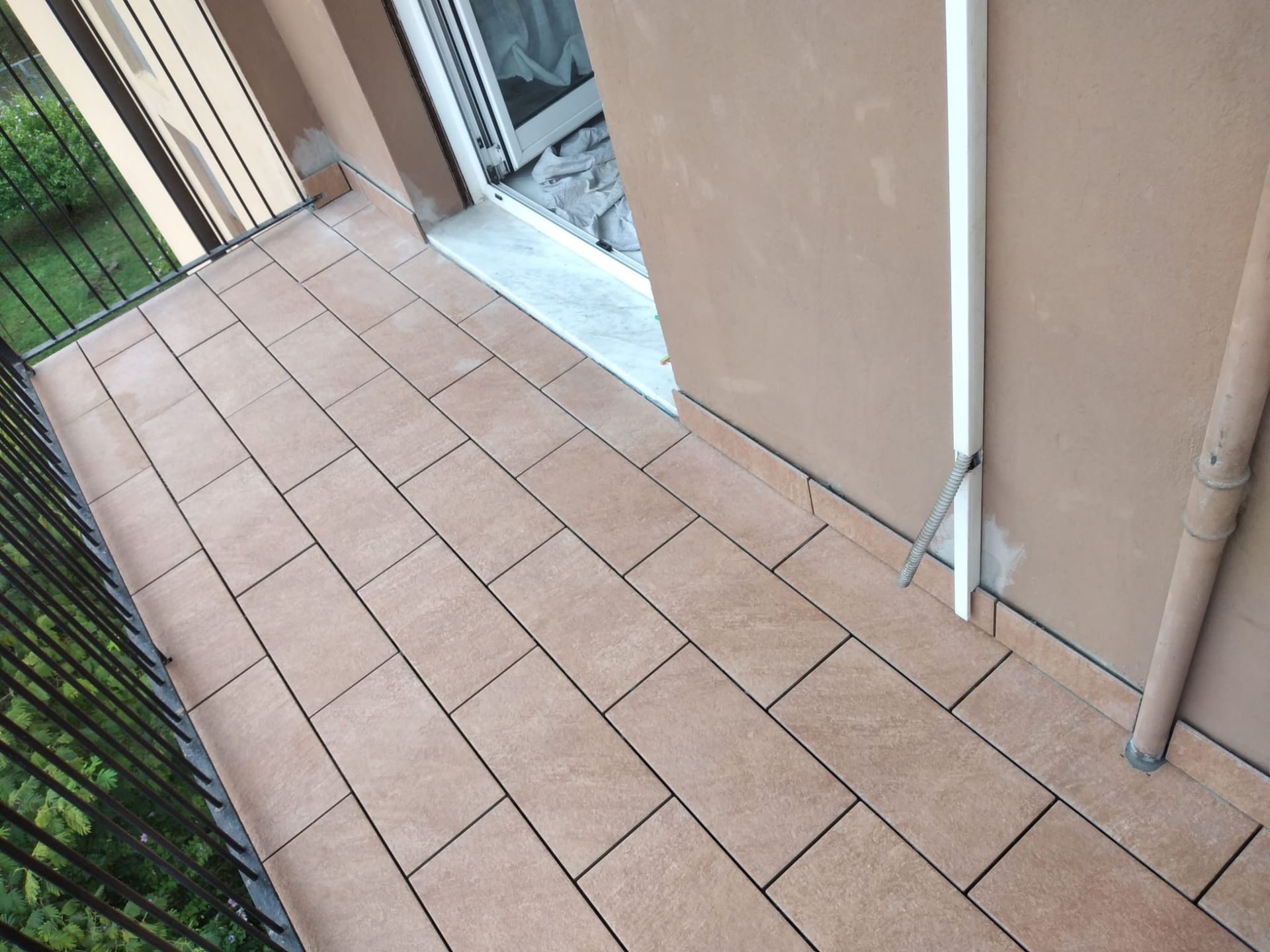 Un balcone con pavimento piastrellato color marrone chiaro, ringhiera in metallo e parete beige con un tubo verticale bianco.
